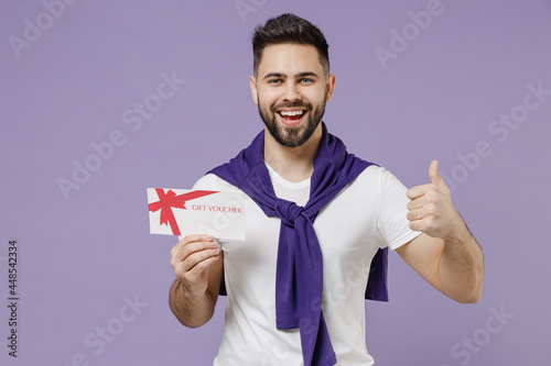 Satisfied fun young brunet man 20s wears white t-shirt purple shirt hold gift certificate coupon voucher card for store show thumb up like gesture isolated on pastel violet background studio portrait © ViDi Studio