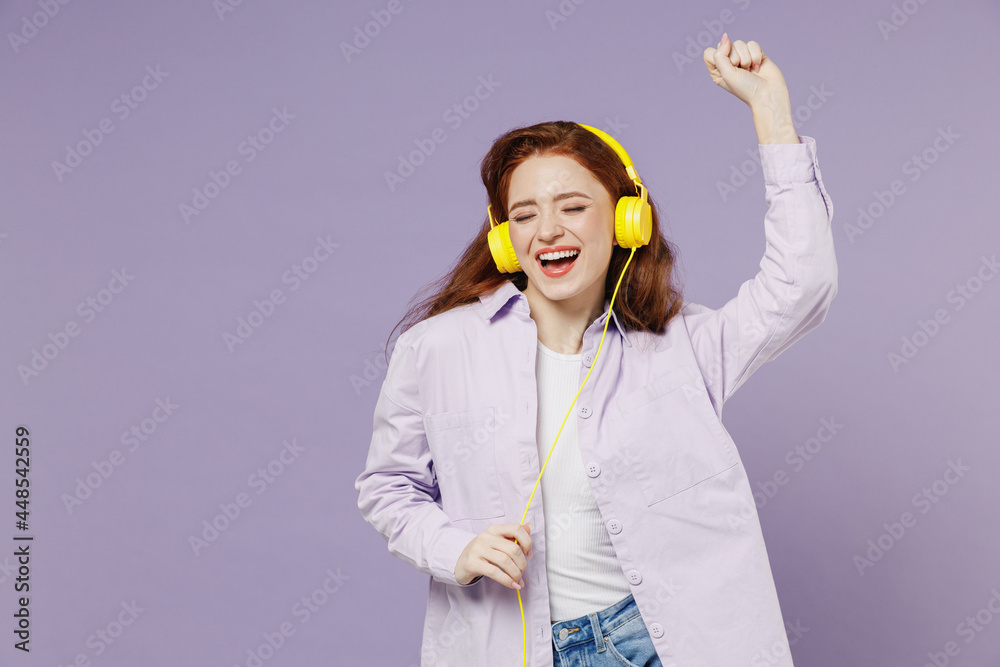 Young redhead curly woman 20s eyes closed mouth open put on white T-shirt violet jacket wear listen to yellow headphones on head dance isolated on pastel purple color wall background studio portrait. Young redhead curly woman 20s eyes closed mouth open put on white T-shirt violet jacket wear listen to yellow headphones on head dance isolated on pastel purple color wall background studio portrait.