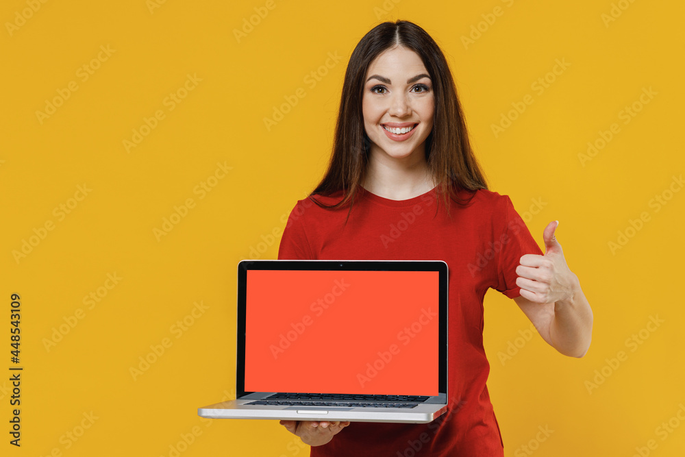 Naklejka premium Smiling young brunette woman 20s wears basic red t-shirt hold use work on laptop pc computer with blank screen workspace area show thumb up like gesture isolated on yellow background studio portrait.