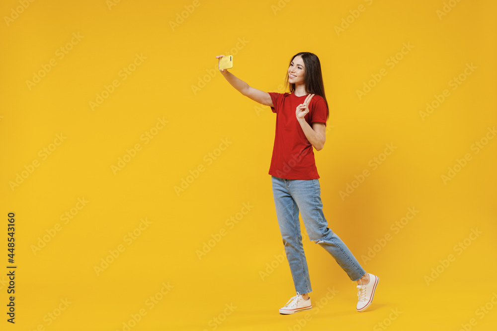 Full size body length young brunette woman 20s wears basic red t-shirt do selfie shot on mobile cell phone post photo on social network show victory sign isolated on yellow background studio portrait