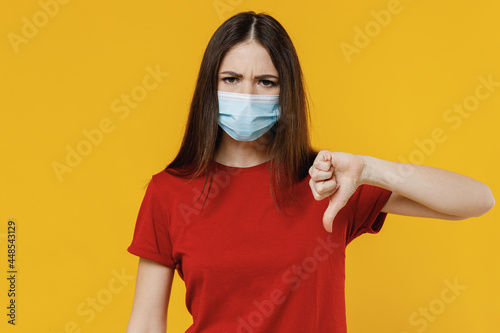 Young brunette woman 20s wear basic red t-shirt sterile face mask ppe to safe from coronavirus virus covid-19 on lockdown show thumb down dislike gesture isolated on yellow background studio portrait