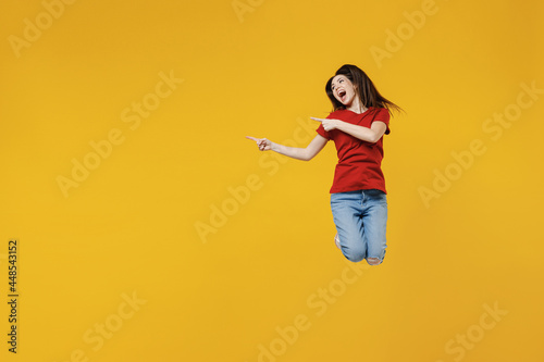 Full size body length young brunette woman 20s wear basic red t-shirt jump point aside away on workspace area copy space mock up isolated on yellow background studio portrait. People emotions concept