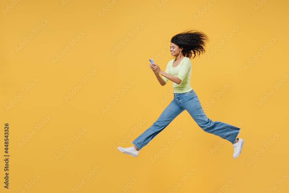 Full size body length side view overjoyed happy african american young woman 20 wear green shirt jump hold in hand use mobile cell phone keep eyes closed isolated on yellow background studio portrait
