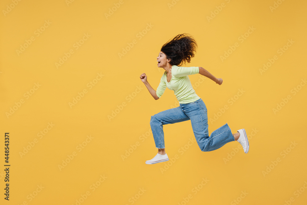 Full size body length side view smiling happy african american young woman 20s wears green shirt jump running closed isolated on yellow background studio portrait. People emotions lifestyle concept