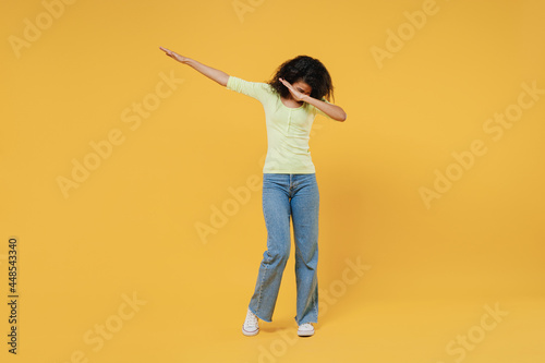 Full size body length slim sporty african american young woman 20s wears green shirt doing dab hip hop dance hands move gesture youth sign hide cover faceisolated on yellow background studio portrait