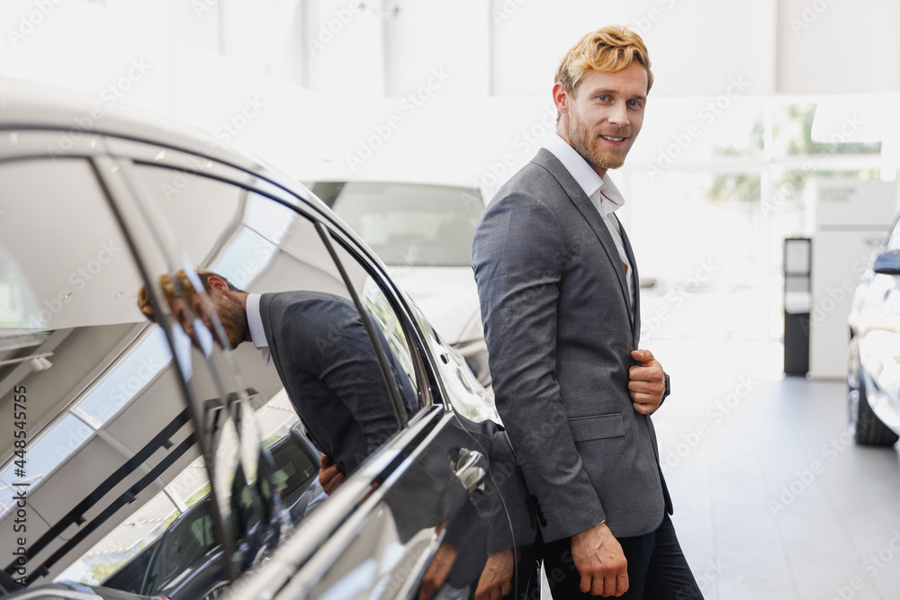 Side view man customer businessman male buyer client in classic grey suit lean on car choose auto wants to buy new automobile in showroom vehicle salon dealership store motor show indoor Sale concept