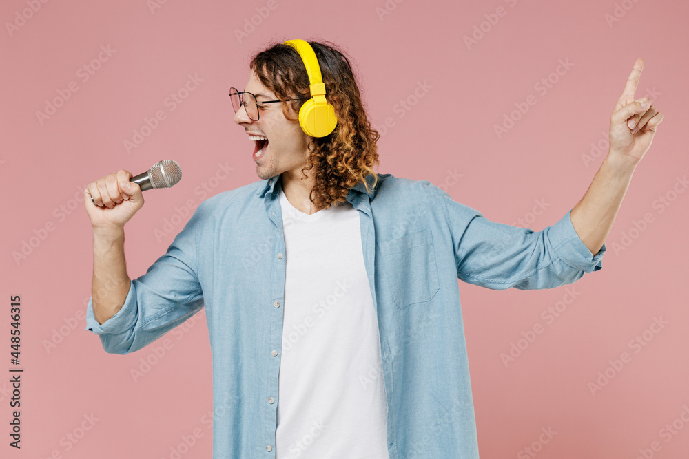 Young singer expressive man 20s with long curly hair wearing blue shirt white t-shirt glasses headphones sing song in microphone isolated on pastel plain pink color wall background studio portrait Young singer expressive man 20s with long curly hair wearing blue shirt white t-shirt glasses headphones sing song in microphone isolated on pastel plain pink color wall background studio portrait