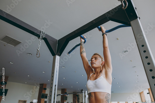Bottom view young skinny caucasian strong sporty athletic sportswoman woman 20s wear white sportswear warm up training pulls up on horizontal bar on in gym indoors Workout sport motivation concept