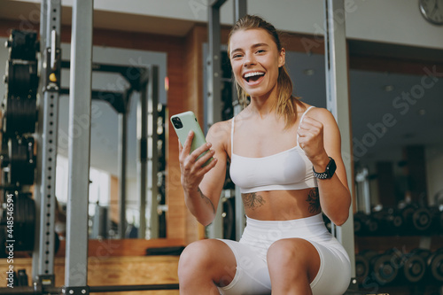 Young overjoyed strong sporty athletic sportswoman woman in white sportswear warm up training sitting near treadmill trainers using mobile cell phone in gym indoors Workout sport motivation concept