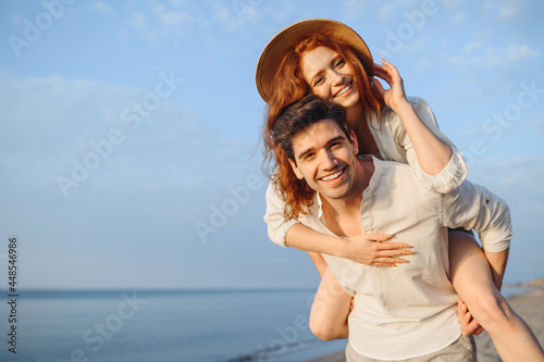 Young cheerful happy couple two friends family man woman in white clothes boyfriend give piggyback ride to joyful, girlfriend sit on back at sunrise over sea beach ocean outside seaside in summer day