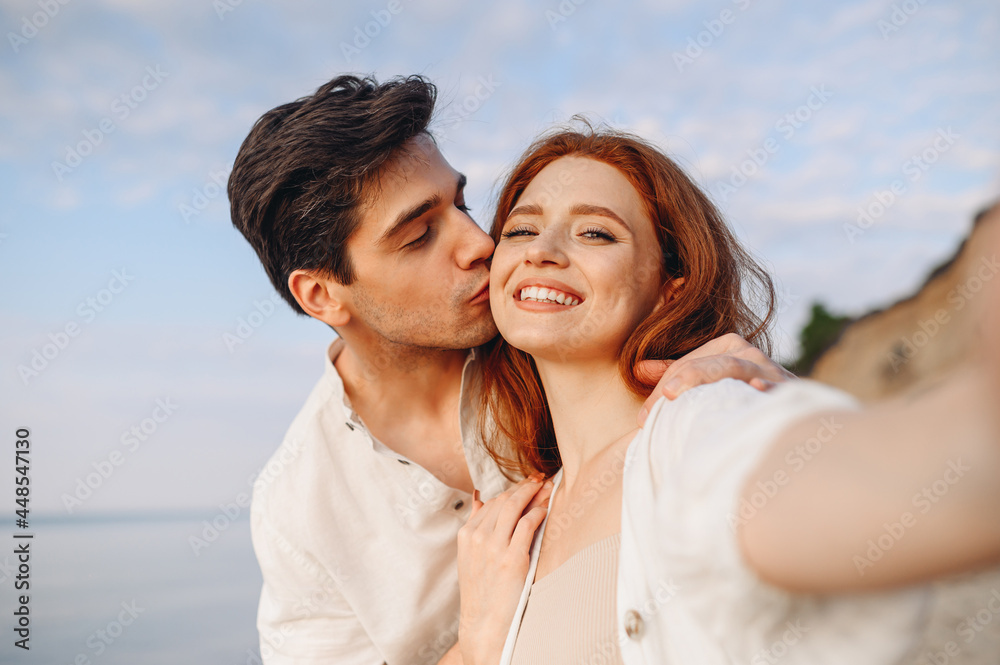Close up happy tender young couple two family man woman in casual clothes hug each other do selfie shot pov on mobile phone kiss at sunrise over sea beach outdoor seaside in summer day sunset evening. Close up happy tender young couple two family man woman in casual clothes hug each other do selfie shot pov on mobile phone kiss at sunrise over sea beach outdoor seaside in summer day sunset evening.