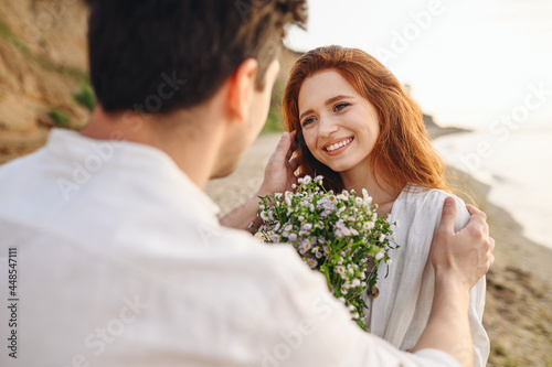 Thankful young smiling couple family man woman in white clothes rest relax together boyfriend meet girlfriend gift give bouquet flowers at sunrise over sea sand beach outdoor seaside in summer day.