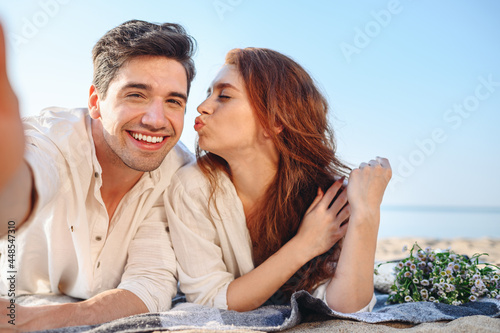 Close up young couple two family man woman in white clothes lying on plaid do selfie shot pov on mobile phone kiss rest together at sunrise over sea beach ocean outdoor seaside in summer day sunset