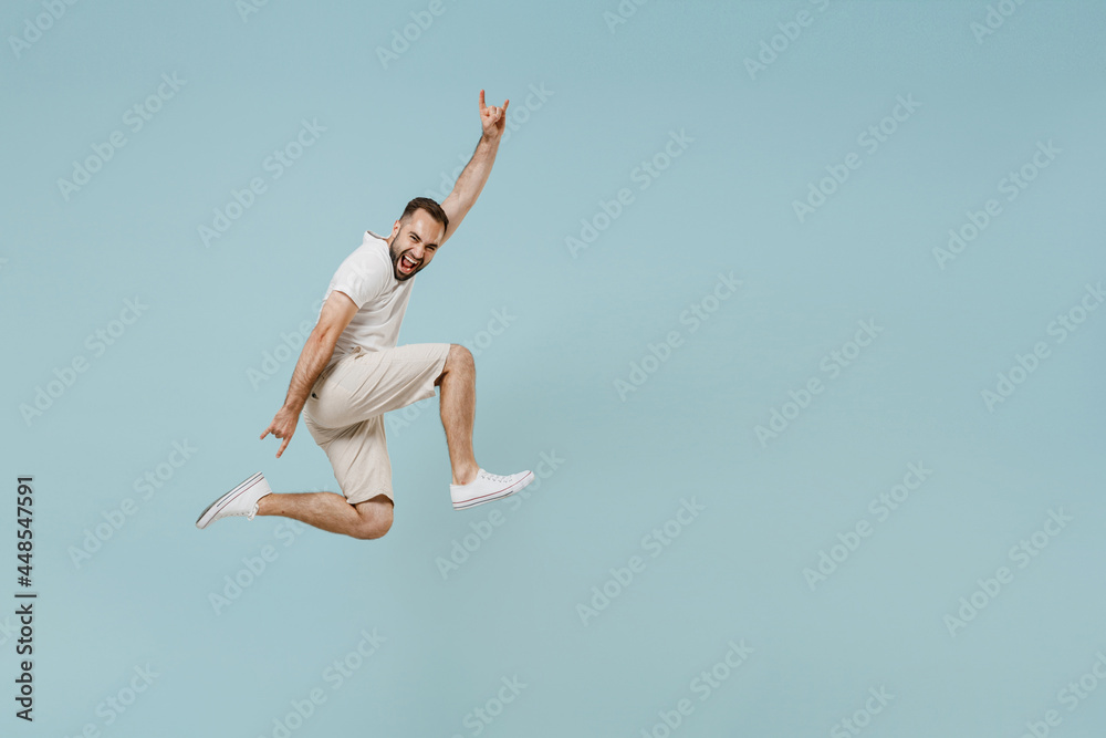 Full length side view young man in white t-shirt jump high horns up gesture, depicting heavy metal rock sign, rock-n-roll, fooling isolated on plain pastel light blue color background studio portrait Full length side view young man in white t-shirt jump high horns up gesture, depicting heavy metal rock sign, rock-n-roll, fooling isolated on plain pastel light blue color background studio portrait