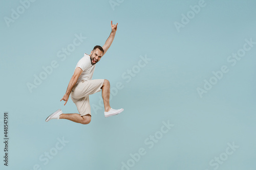 Full length side view young man in white t-shirt jump high horns up gesture, depicting heavy metal rock sign, rock-n-roll, fooling isolated on plain pastel light blue color background studio portrait