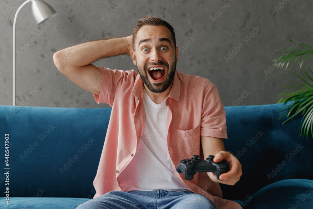 Young excited surprised man 20s in casual clothes hold in hand play pc game with joystick console do winner gesture clench fist sit on blue sofa at home flat indoors rest relax on weekends free time