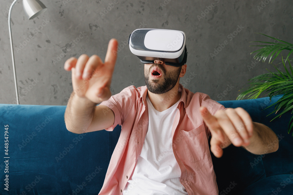 Naklejka premium Young smiling happy man wear casual clothes watching in vr headset pc gadget press button sitting on blue sofa at home flat indoors rest relax on weekends free time. People lounge lifestyle concept