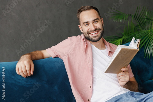 Fotografie Young smiling happy satisfied european man 20s wearing casual clothes reading book novel sitting on blue sofa at home flat indoors rest relax on weekends free time