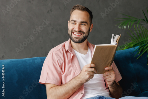 Obraz na plátně Young smiling happy pensive man 20s wearing casual clothes reading book novel look aside sitting on blue sofa at home flat indoors rest relax on weekends free time
