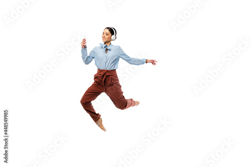 Smiling ballerina in headphones using smartphone while jumping isolated on white