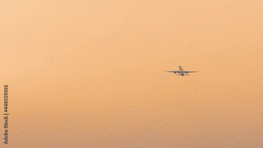 Airplane flies high in the air against orange sky. Sunset or sunrise colors. Aircraft ascending and travelling away. Slow motion view of large passenger plane above Airplane flies high in the air against orange sky. Sunset or sunrise colors. Aircraft ascending and travelling away. Slow motion view of large passenger plane above