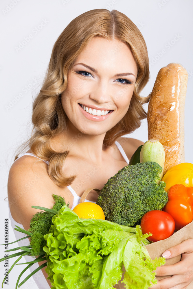 Portrait photo of happy smiling young beautiful woman holding grocery shopping bag with healthy vegetarian raw food, isolated on gray background. Attractive blond girl at studio. Portrait photo of happy smiling young beautiful woman holding grocery shopping bag with healthy vegetarian raw food, isolated on gray background. Attractive blond girl at studio.