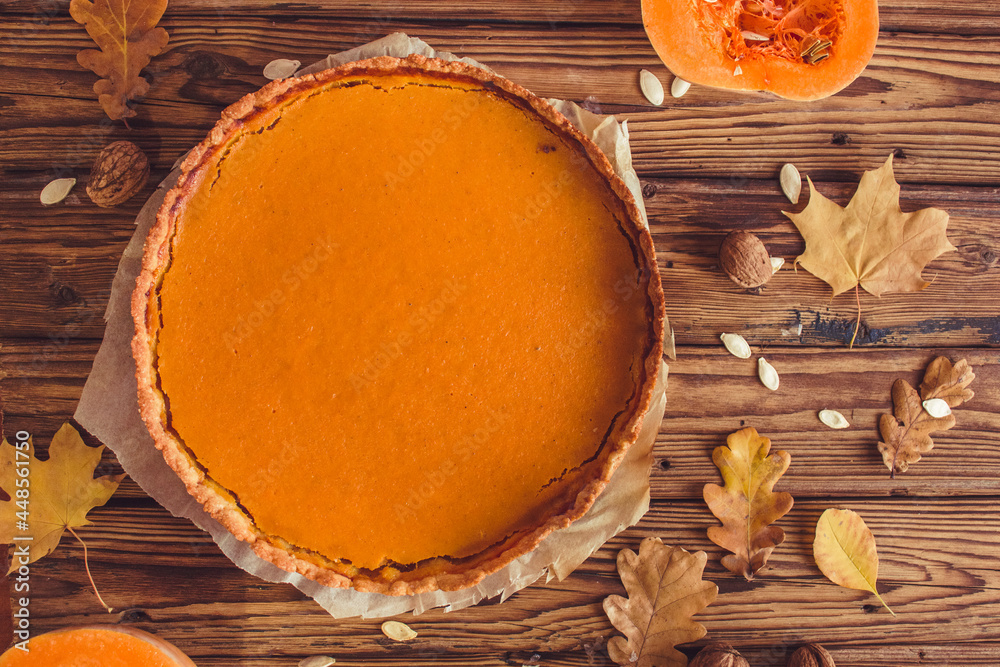 Orange spicy autumn pumpkin pie. Shortcrust vanilla tart with sweet pumpkin cream on brown wooden background with small sliced pumpkins walnuts oak leaves and seeds. Free copy space. Top view Orange spicy autumn pumpkin pie. Shortcrust vanilla tart with sweet pumpkin cream on brown wooden background with small sliced pumpkins walnuts oak leaves and seeds. Free copy space. Top view