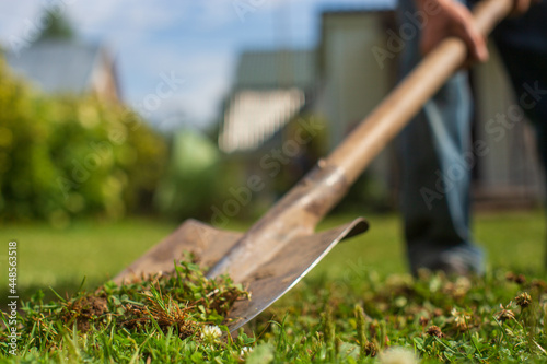 A farmer with a shovel digs the ground in the garden. The shovel blade is in the foreground and goes into the bokeh zone. In the foreground is green grass.