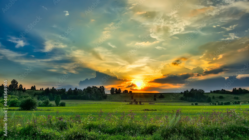 Obraz premium Summer sunset over green field - amazing rural landscape. Golden sunbeams illuminating clouds, hills and valley with wheat field. Colorful wildflowers at foreground. Beauty of summer nature