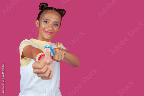 Young latina girl plays and points at you with a hair garter on a colorful background while smiling. Horizontal photo
