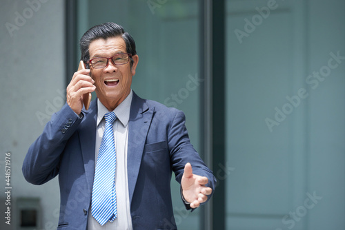Excited aged businessman standing outside the office building and talking on phone with customer