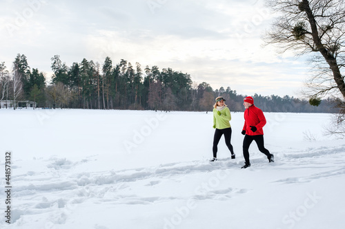 Smiling senior couple jogging in snowy winter park. Elderly wife and husband doing workout outdoors. Active lifestyle concept.