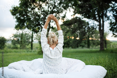 Young blonde woman wake up wear pajamas sitting in bed on white blanket outdoors over green nature background. Good morning. Back view. Relaxation. Summer vacation season. #448573581