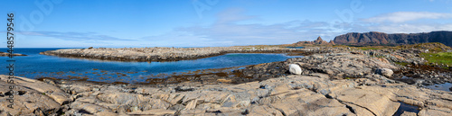 Wide panorama. Rocky stone coast on the shore of a blue fjord with an island on the horizon under a blue sky. A mountain range with a lonely peak. Leka Island. Norway.