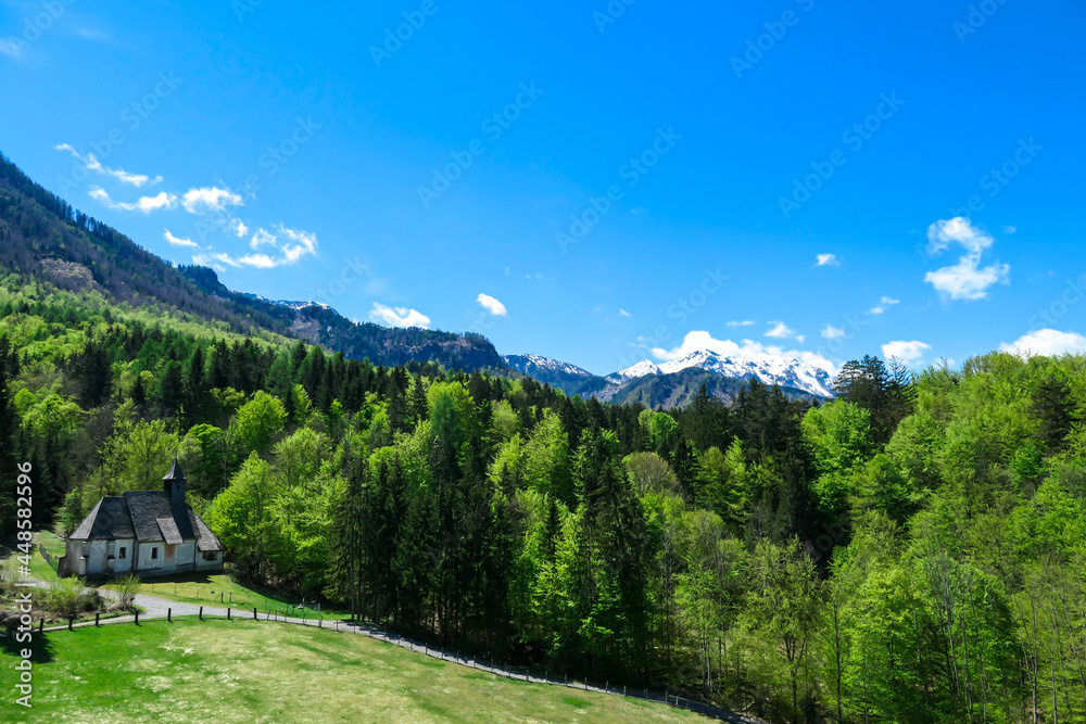A a small chapel at the edge of the forest the panoramic view on Baeren Valley in Austrian Alps. The highest peaks are snow-capped. Lush green pasture. Clear and sunny day. High mountain chains.