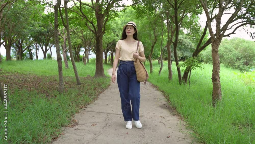 full length asian female traveler wearing a hat is looking around while taking a stroll alone on the path in the woods on summer’s day at dapo lake, taitung.