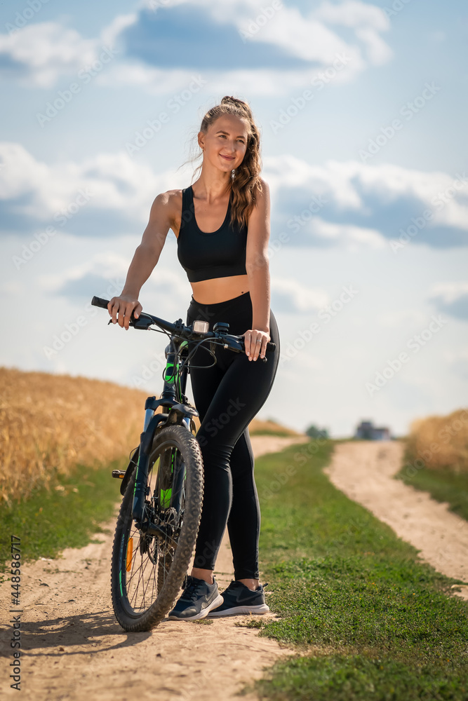 Young athletic woman in leggings and a top with a bicycle stands on the road in the middle of a field with triticale on a sunny summer day. Vertical photo Young athletic woman in leggings and a top with a bicycle stands on the road in the middle of a field with triticale on a sunny summer day. Vertical photo