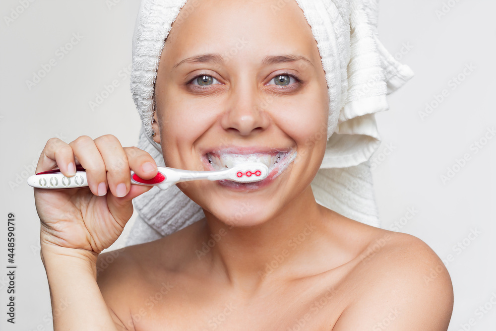A young beautiful caucasian smiling woman with a white towel on her head after a shower is brushing her teeth with a toothbrush on a white background. Morning and evening routine. Close up A young beautiful caucasian smiling woman with a white towel on her head after a shower is brushing her teeth with a toothbrush on a white background. Morning and evening routine. Close up