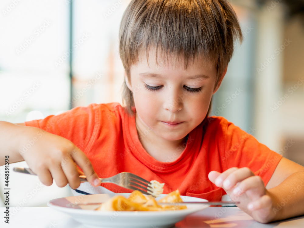 Cute kid in red t-shirt eats pancakes with knife and fork. Curious boy with funny expression on face. Tasty pastry for breakfast. Cute kid in red t-shirt eats pancakes with knife and fork. Curious boy with funny expression on face. Tasty pastry for breakfast.
