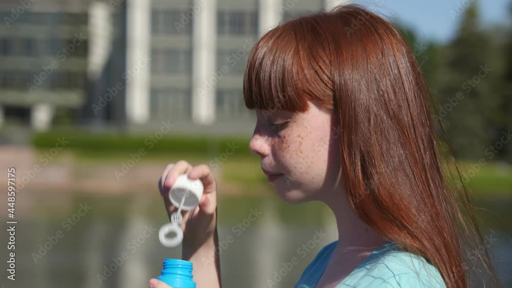 Side view of teenage redhead girl blowing soap bubbles outdoors.