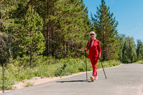 an elderly woman in red tracksuit walks with Nordic walking sticks in the summer