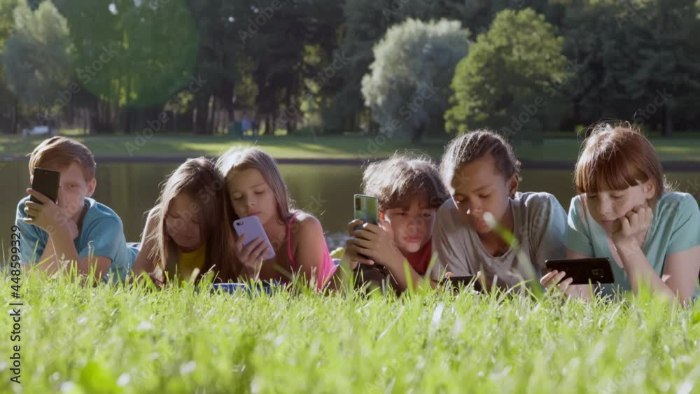 Multiethnic teen children using mobile phone lying together on grass in spring park Multiethnic teen children using mobile phone lying together on grass in spring park