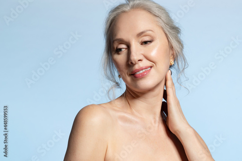Beautiful 50s gray-haired senior female with naked shoulder and perfect groomed skin, studio portrait on blue with copy space. Attractive mature lady touching neck. Beauty and aged skincare concept