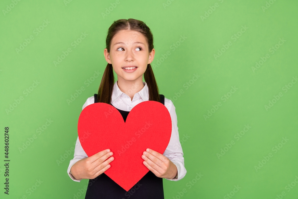 Photo of young school girl happy positive smile hold big red paper heart dream look empty space isolated over green color background Photo of young school girl happy positive smile hold big red paper heart dream look empty space isolated over green color background