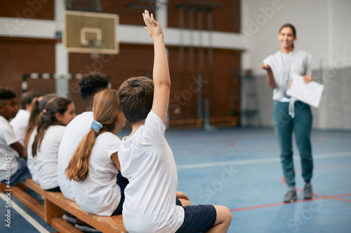 Rear view of schoolboy raising hand to ask question during physical education class.