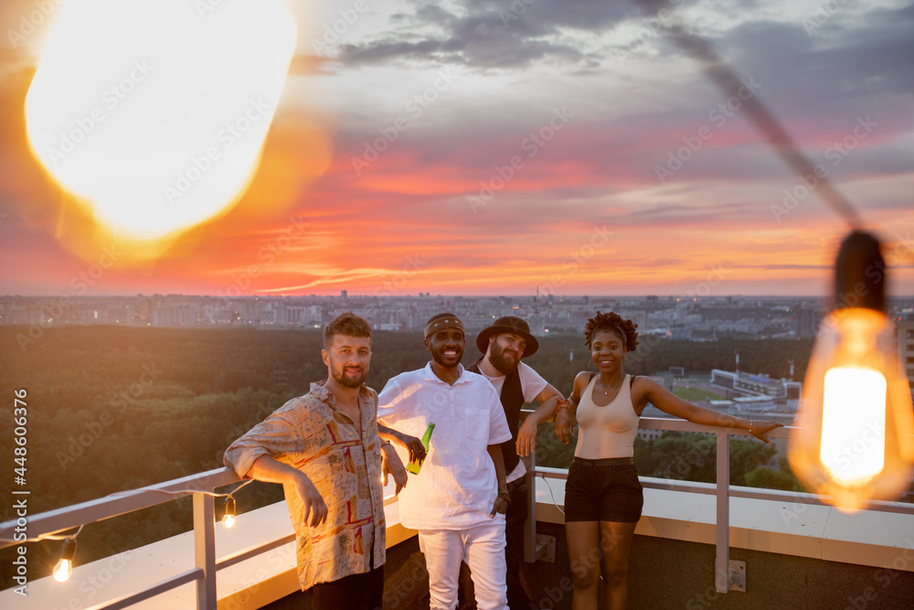 Fototapeta premium Four happy young friends standing on rooftop patio against cityscape and cloudy sky