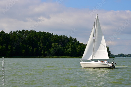 View of small boat or a passenger vessel alike with a white sail and a light body sailing slowly through a vast yet shallow river or lake with some forest or moor growing near to a reed covered coast