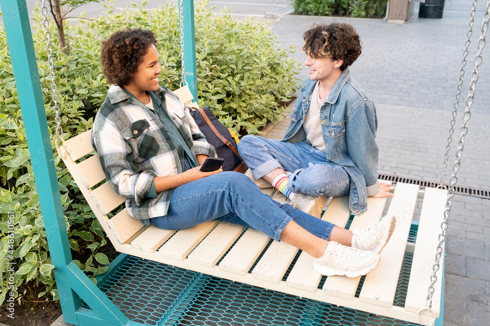 Teenage romantic dates in casualwear chatting on wooden swings outdoors