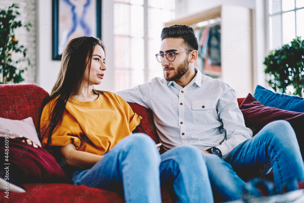 Turkish boyfriend in spectacles talking with girlfriend discussing plans on weekend while resting at comfortable sofa, couple in love spending leisure time together talking on couch in stylish room