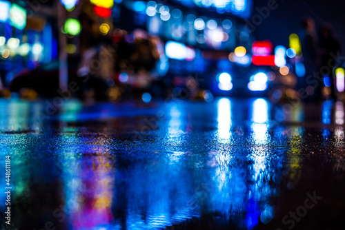 Rainy night in the big city, light from the shop windows reflected on the road on which cars travel. View from the level of asphalt